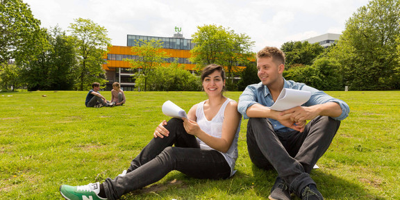 Zwei Studierende sitzen auf einer Wiese, im Hintergrund die Mensa