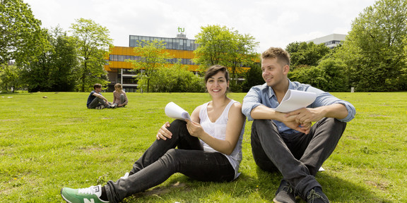 Zwei Studierende sitzen auf einer Wiese, im Hintergrund die Mensa